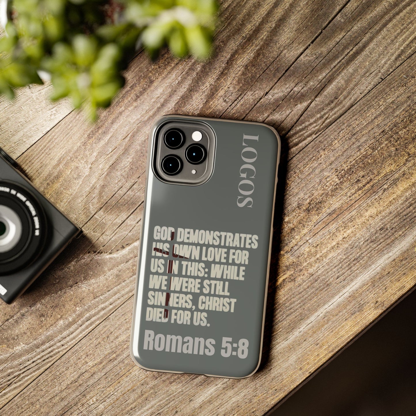 Logos Gray Christian phone case with Romans 5:8 scripture text on a wooden surface