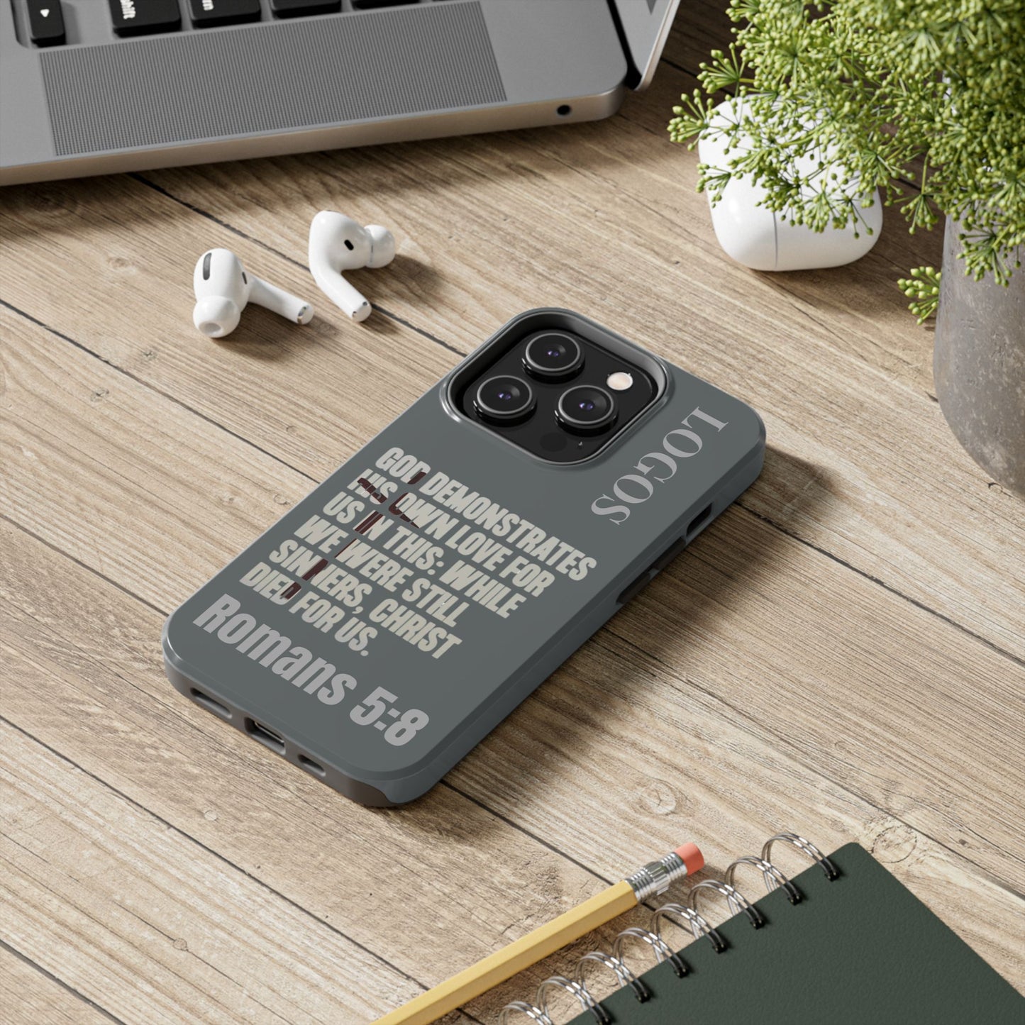Logos Christian phone case with Bible verse Romans 5:8 in gray on a wooden desk