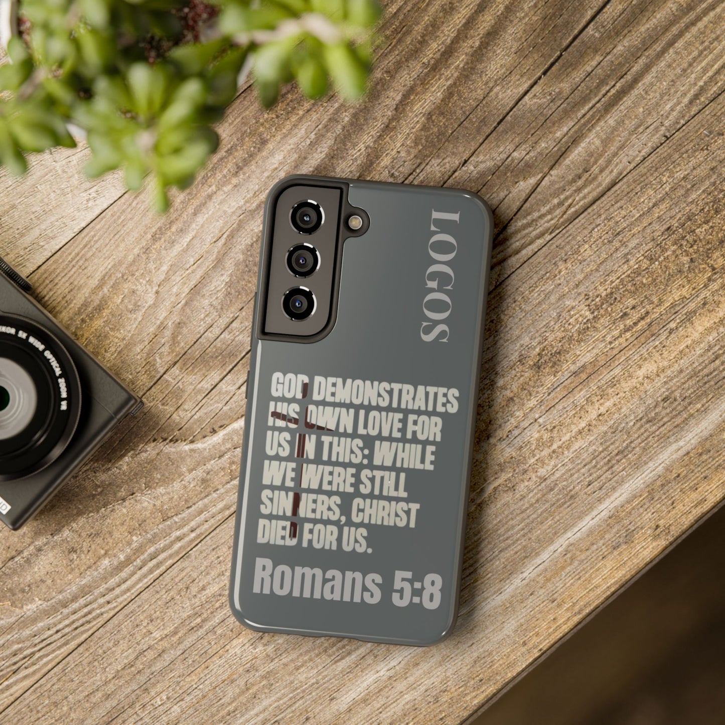 Logos Christian phone case in gray with Bible verse Romans 5:8 on a wooden surface
