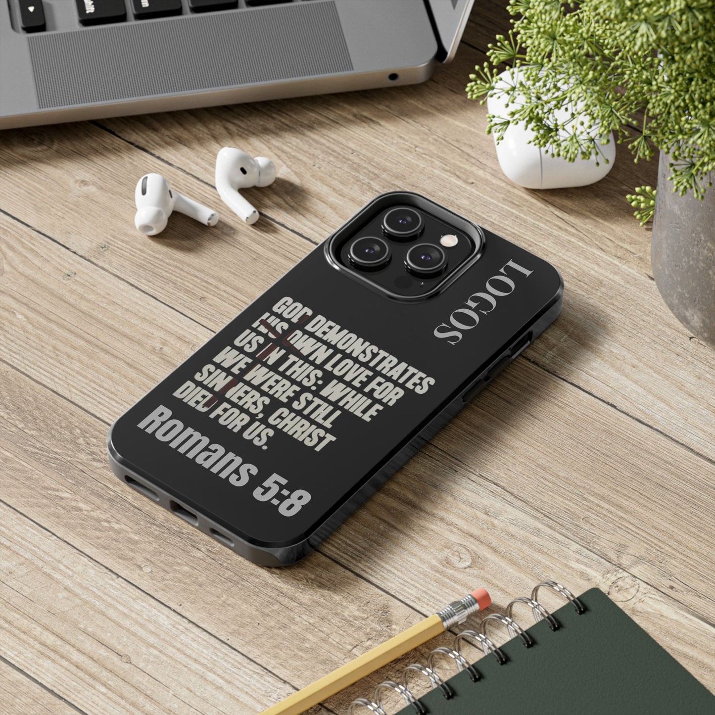 Romans 5:8 phone case by Logos with black design and inspirational Bible verse text on wooden desk