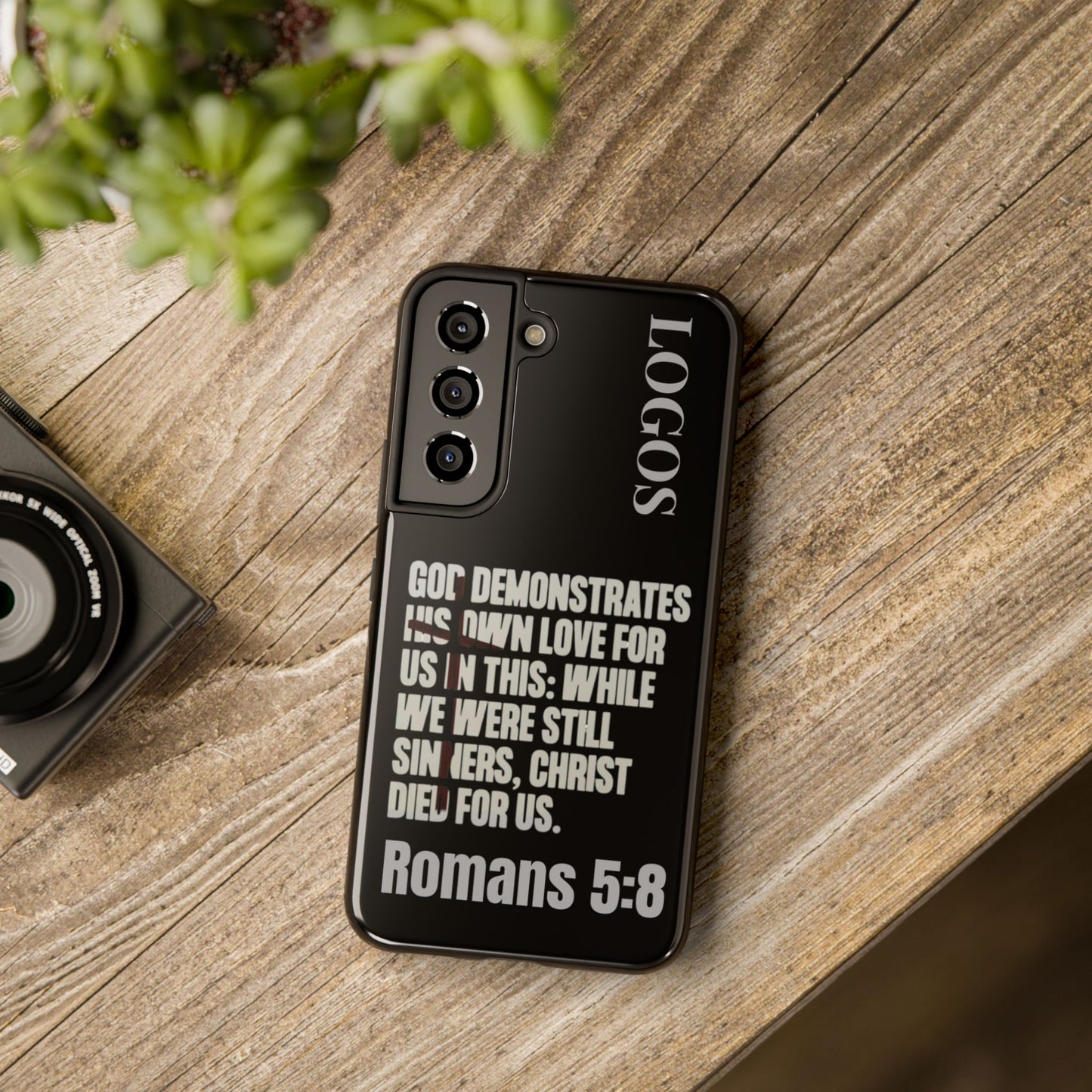 Black phone case with Bible verse Romans 5:8 text design by Logos on wooden surface