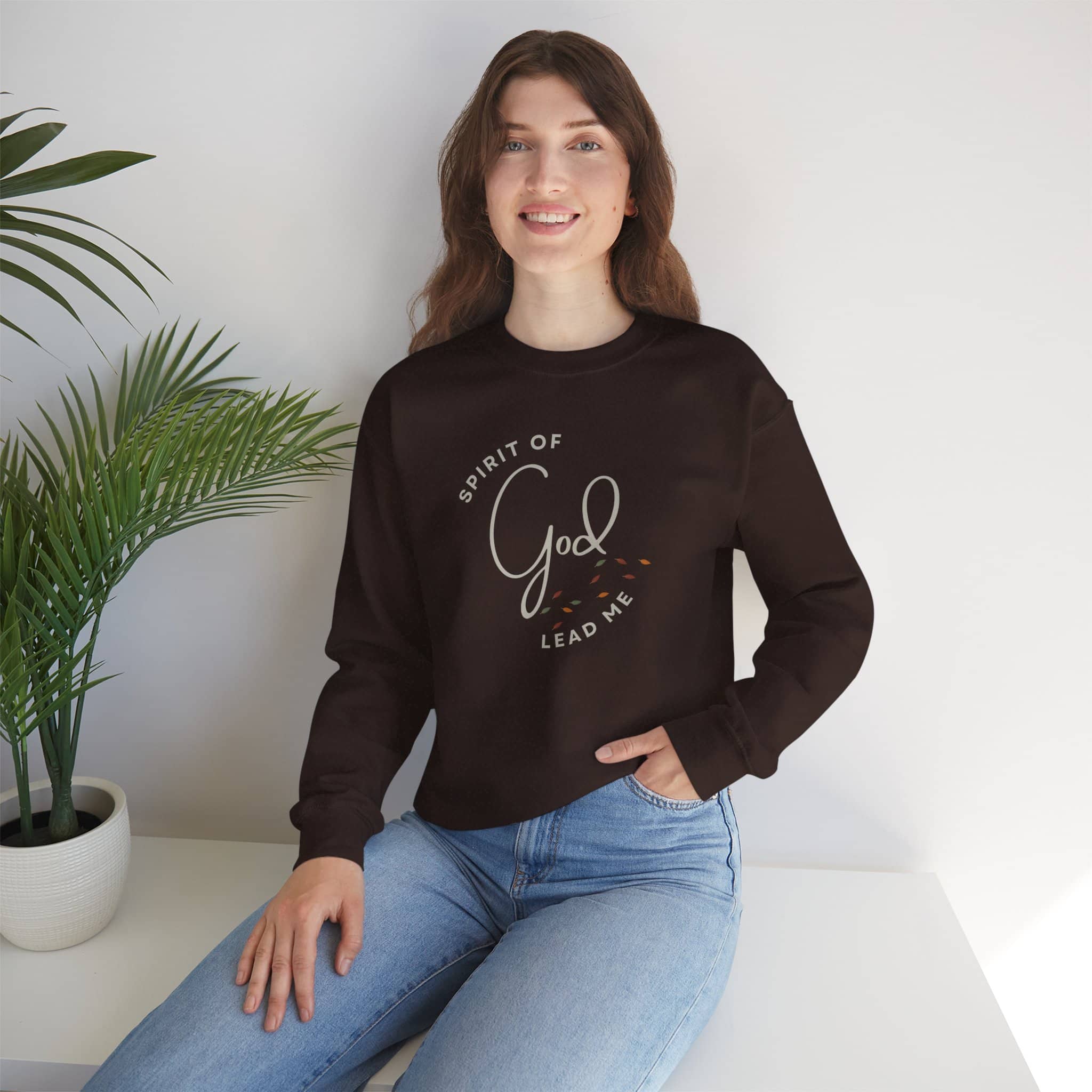 Woman wearing black Spirit Lead Me sweatshirt with God script design sitting by potted plant