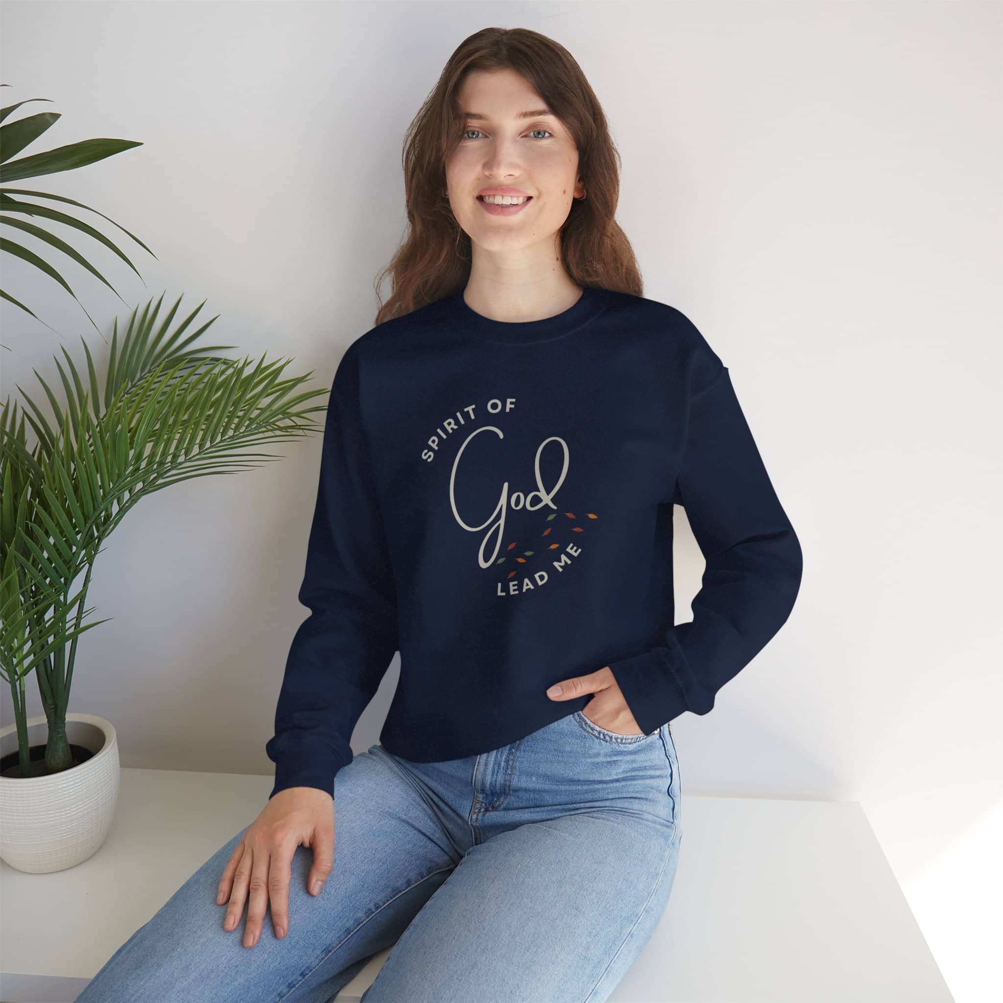 Woman wearing navy blue Spirit Lead Me sweatshirt with Spirit of God lead me text design sitting by a plant